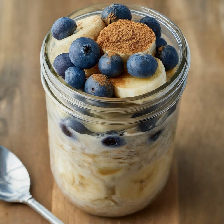 A glass jar filled with Blueberry-Banana Overnight Oats layered with rolled oats, mashed banana slices, and fresh blueberries, topped with a sprinkle of cinnamon and a spoon beside the jar.