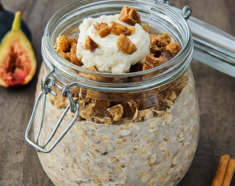 A close-up photo of a glass jar filled with layered Fig & Ricotta Overnight Oats. Rolled oats, chopped dried figs, and ricotta cheese are visible, with a cinnamon stick and spoon beside the jar.