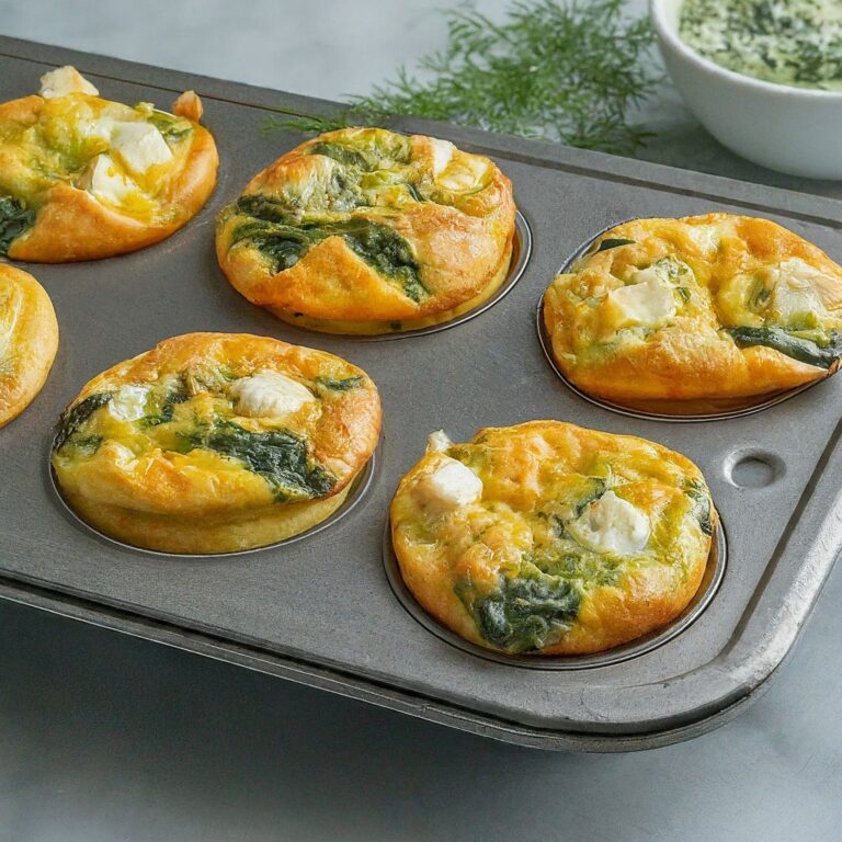 A close-up view of a muffin tin containing freshly baked Spanakopita Egg Muffins. The golden brown muffins are slightly puffed and filled with spinach and feta cheese. A small ramekin filled with the spinach filling rests beside the muffin tin, accompanied by a sprig of fresh dill for garnish.