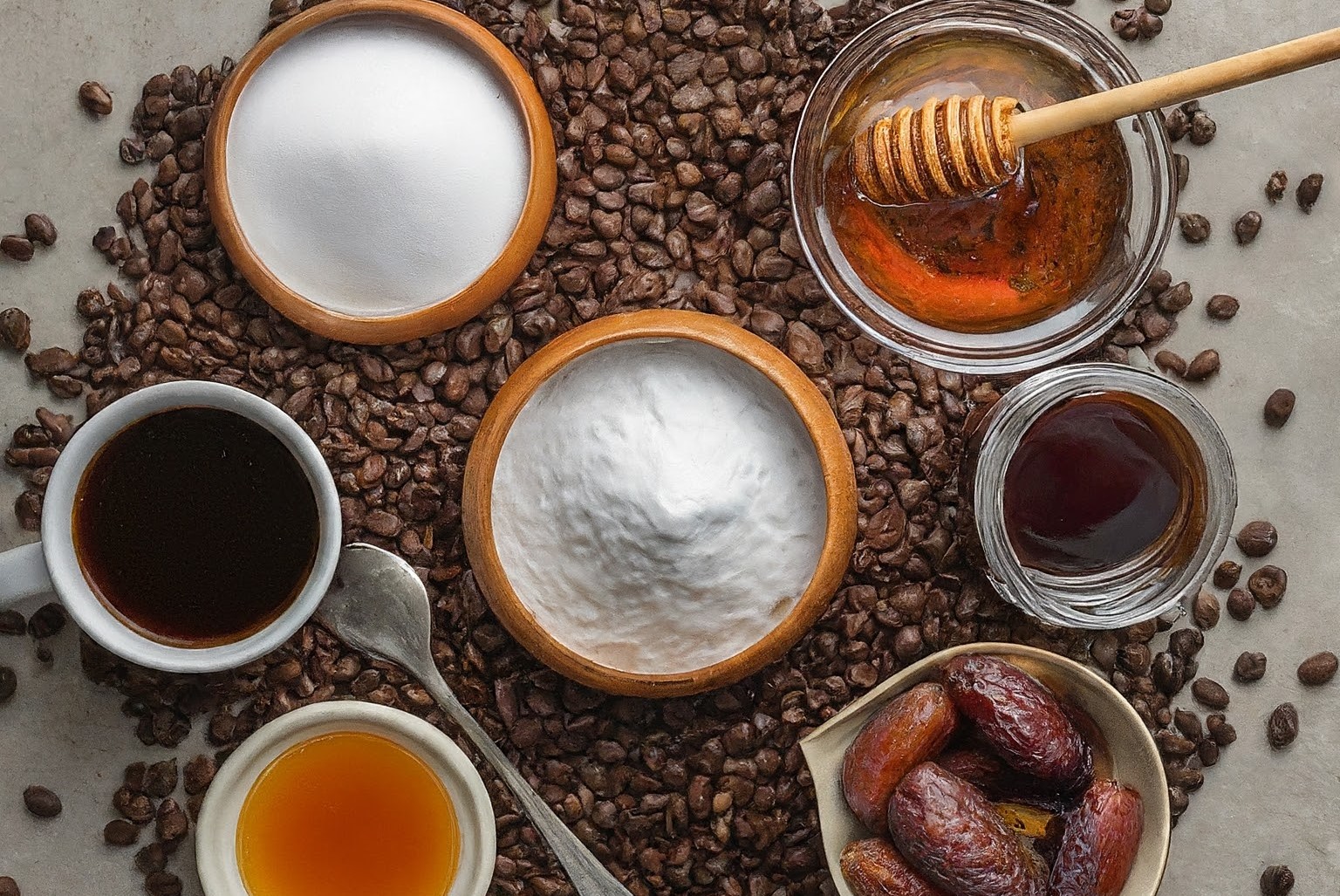 Flat lay of natural coffee sweeteners. Stevia, monk fruit, erythritol, xylitol, coconut sugar, honey, maple syrup, date syrup, coffee beans, and a steaming cup of coffee.