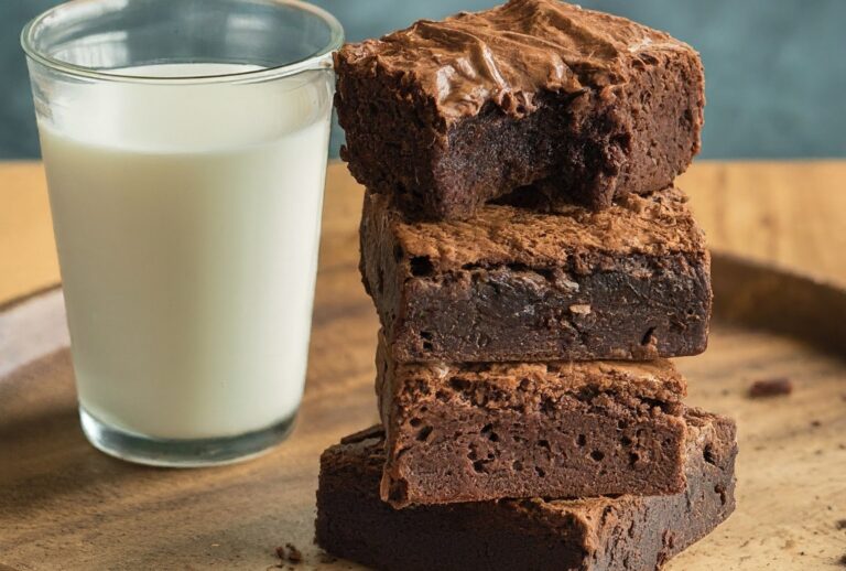 A split image with two halves. The left half showcases a classic fudgy brownie with a rich, dark chocolate color and a slightly cracked top. Next to it, a more cakey brownie sits with a lighter chocolate color and a smooth, even top. Both brownies have a single bite taken out, revealing their moist and delicious interiors. In the background, a glass of milk sits on the left next to the fudgy brownie, and a glass of water sits on the right next to the cakey brownie. The text "Milk vs. Water: The Great Brownie Debate" sits above the image.