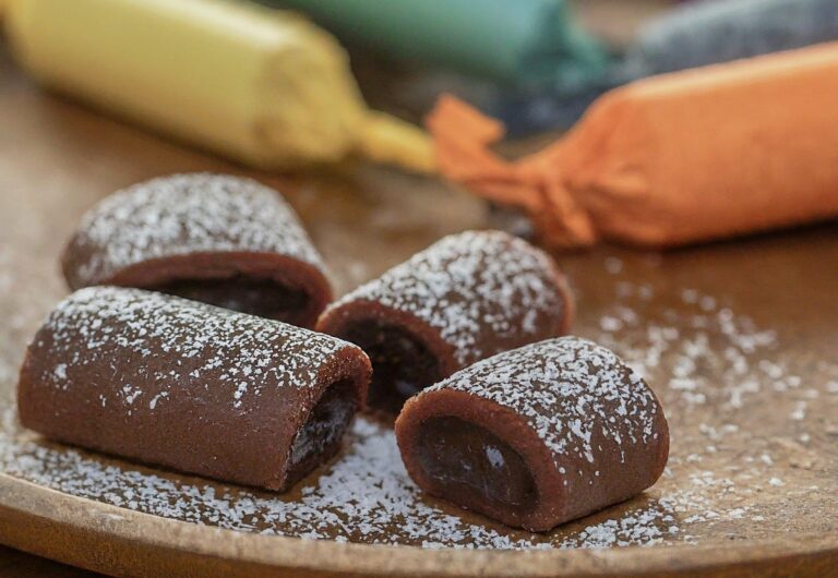 A close-up shot of several homemade Tootsie Rolls arranged on a plate. Some Tootsie Rolls can be unwrapped, revealing the rich chocolatey center, while others remain wrapped in colorful wrappers (you can use colored parchment paper for this). A sprinkle of powdered sugar can be dusted around the Tootsie Rolls for added visual interest.